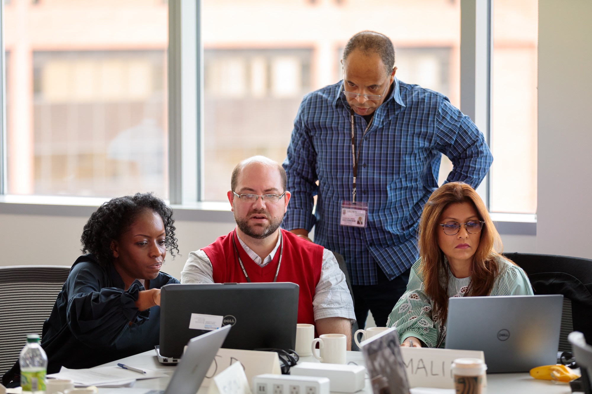 Four coworkers collaborate around a conference table with laptops in a bright office.