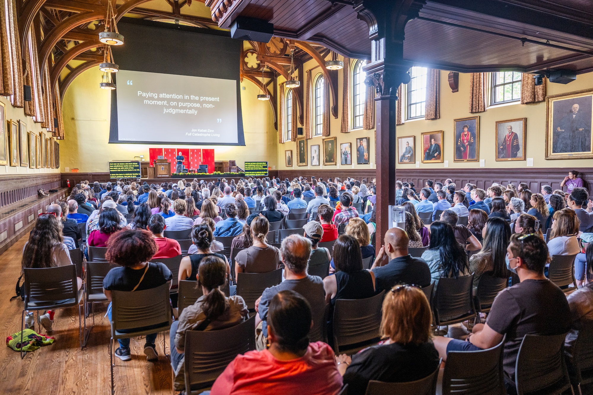 Wide shot from the back of a large, wood-paneled hall filled with an audience listening to a speaker on stage. A large projection screen displays a quote by Jon Kabat-Zinn about mindfulness.